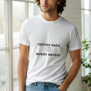 Op de afbeelding: Wit T-shirt met ronde hals met de tekst "COFFEE DATA QUERY REPEAT" in zwart. Het shirt heeft een koffiekopje-icoon. Het model draagt een blauwe spijkerbroek. Het shirt is gemaakt van een zacht, comfortabel materiaal.