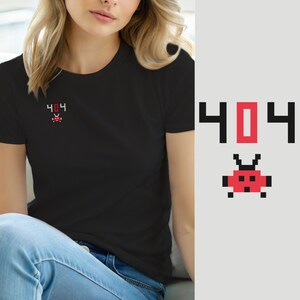 Puede incluir: Una camiseta negra de manga corta con un pequeño gráfico de pixel art en el pecho. El diseño consiste en el texto 404 en negro y rojo, situado sobre una criatura alienígena pixelada roja y negra con dos antenas y un cuerpo cuadrado.