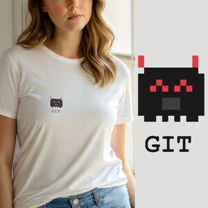 Puede incluir: Una camiseta blanca de manga corta con un pequeño gráfico pixelado de una criatura negra con ojos rojos y dos antenas rojas. Debajo de la criatura, el texto GIT está impreso en una fuente negra sencilla sin remates en el área del pecho.