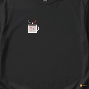 Puede incluir: Camiseta negra con un diseño pixelado de taza de café. El gráfico incluye una taza blanca con la parte superior marrón, un arco rojo y blanco y un pequeño diseño abstracto dentro de la taza. La camiseta es de algodón pesado.