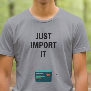 Op de afbeelding: Lichtgrijs t-shirt met de zwarte tekst "JUST IMPORT IT". Daaronder staat een turquoise afbeelding van een doos met lijnen en een pijl. Het shirt met korte mouwen wordt gedragen tegen een wazige groen-bruine achtergrond.