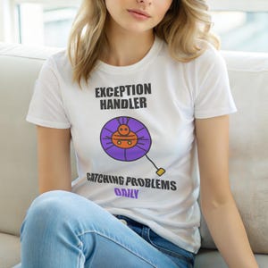 Op de afbeelding: Wit T-shirt met de tekst "EXCEPTION HANDLER" boven een paarse en oranje afbeelding. Daaronder staat "CATCHING PROBLEMS DAILY". Het shirt wordt gecombineerd met een blauwe jeans.