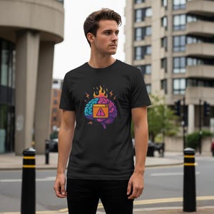 May include: Black t-shirt featuring a colorful brain graphic with a warning symbol. The brain design includes flames, lightning bolts, and a gradient of blue, orange, and purple. The t-shirt is worn by a person standing outdoors.