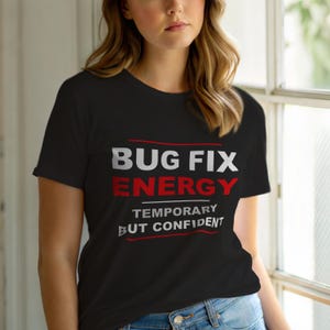Op de afbeelding: Zwart t-shirt met de tekst "BUG FIX ENERGY" in wit en rood, met daaronder de woorden "TEMPORARY BUT CONFIDENT". Het shirt heeft korte mouwen en lijkt van een zacht materiaal te zijn.