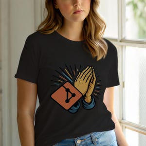 May include: Black t-shirt with a graphic of praying hands and a geometric design on a square. The hands are tan, the square is orange, and the design is black. The shirt has short sleeves.