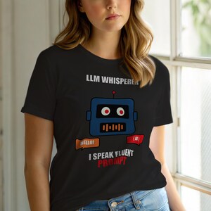 以下が含まれることがあります： 黒いTシャツ。青いロボットのデザインと「LLM Whisperer」の文字。ロボットは赤い目とスピーカーのグラフィック。下に「I Speak Fluent Prompt」と「Hello!」と「{AI}」の吹き出しがあります。