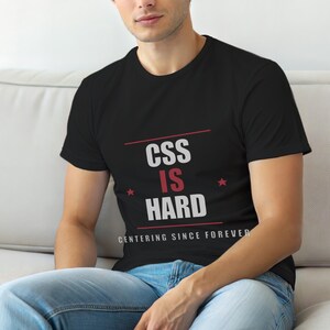 Op de afbeelding: Zwart T-shirt met de tekst "CSS IS HARD" in wit en rood, met de zin "CENTERING SINCE FOREVER" eronder. Het shirt heeft twee rode sterren. De persoon draagt een blauwe spijkerbroek.