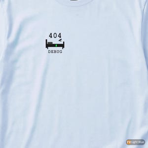 May include: Light blue t-shirt with a black graphic featuring the number "404", a bed, and the word "DEBUG" in black. The bed has a green bar in the middle. The shirt is made of a soft material.