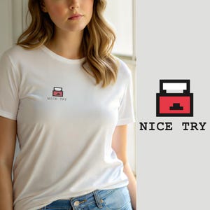 Op de afbeelding: Een wit T-shirt met korte mouwen en een kleine, gepixelde afbeelding op de linkerborst. De afbeelding toont een rode rechthoekige doos met een witte bovenkant en zwarte omlijningen, vergezeld van de zwarte tekst NICE TRY eronder.