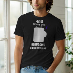 May include: Black t-shirt with a white graphic of a coffee mug and the text "404 Sleep Not Found" and "Debugging Since Midnight". The shirt is a crew neck and is being worn by a person.