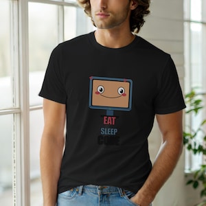 Puede incluir: Camiseta negra con un diseño de monitor de ordenador de dibujos animados sonriente. El texto "EAT SLEEP CODE" está impreso debajo. El monitor tiene mejillas rosas y acentos de corazón rojo.