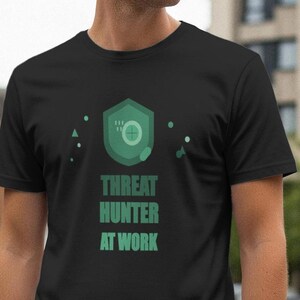 May include: Black t-shirt featuring a green shield design with a circle and other shapes, and the text "THREAT HUNTER AT WORK". The shirt is made of a soft material.