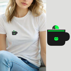 Pode incluir: Uma t-shirt branca de manga curta com um pequeno gráfico no peito. O gráfico mostra uma caneca de café preta com uma borda verde brilhante e uma pequena criatura verde estilizada espreitando por cima, com um ícone verde correspondente.
