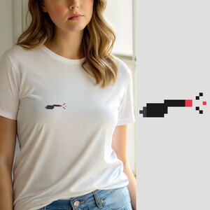 Op de afbeelding: Een wit T-shirt met korte mouwen en een kleine pixel art afbeelding op de borst. De afbeelding toont een donkergrijs en rood object dat lijkt op een spuitmond, met kleine gepixelde stippen die uit de punt komen om spray weer te geven.
