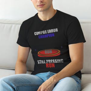Może przedstawiać: Czarna koszulka z napisem "COMPILE ERROR CHAMPION" w kolorze białym i niebieskim. Poniżej czerwono-pomarańczowa grafika ze słowem "RUN". Pod grafiką znajduje się tekst "STILL PRESSING RUN".
