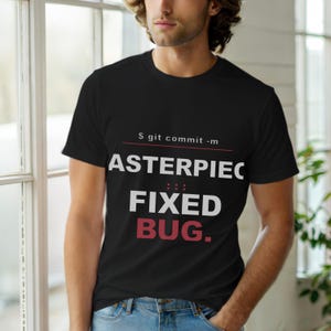 Op de afbeelding: Zwart T-shirt met ronde hals met witte en rode tekst. De tekst luidt "S git commit -m ASTERPIEC FIXED BUG." Het T-shirt wordt gedragen door een persoon voor een raam.