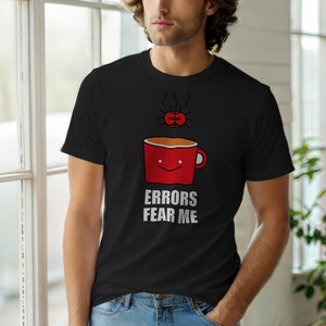 Puede incluir: Camiseta negra con un diseño humorístico. Presenta una taza de café roja con una cara sonriente y el texto "ERRORS FEAR ME" en blanco. Encima de la taza hay un cerebro. La camiseta está hecha de un material suave.