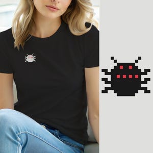 Könnte beinhalten: Ein schwarzes T-Shirt mit einer kleinen, verpixelten Insektengrafik auf der linken Brust. Das Insektendesign besteht aus weißen und roten quadratischen Pixeln und zeigt einen Käfer mit zwei Fühlern, sechs Beinen und einer Reihe roter Punkte.