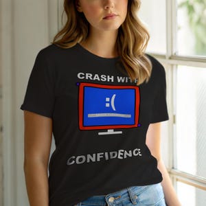Może przedstawiać: Czarny t-shirt z grafiką niebieskiego ekranu komputera z smutną buzią i napisem "Crash With Confidence". Ekran ma czerwoną obwódkę i tekst "Error 0x00000000" pod smutną buzią.