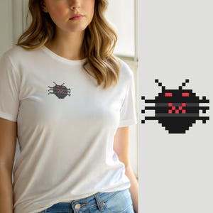 Op de afbeelding: Een wit T-shirt met korte mouwen en een kleine pixel art afbeelding op de linkerborst. De afbeelding toont een zwart rond wezen met zes poten, twee voelsprieten, rode gepixelde ogen en een rode gepixelde mond op een donkergrijze streep.