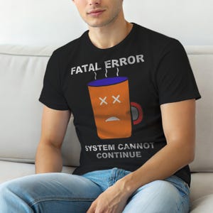 Op de afbeelding: Zwart T-shirt met de tekst "FATAL ERROR" boven een cartoon koffiemok. De mok is oranje met een blauwe rand en rode oor, met doorgestreepte ogen en een verdrietige mond. De tekst "SYSTEM CANNOT CONTINUE" staat eronder.