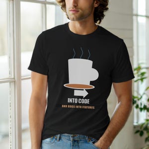 Op de afbeelding: Zwart t-shirt met een witte koffiekop-afbeelding en de tekst "INTO CODE AND BUGS INTO FEATURES". Het ontwerp bevat een bruine schotel en een pijl.