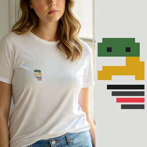 Pode incluir: Uma t-shirt branca de manga curta com um pequeno gráfico pixelizado no peito. O design consiste numa cabeça retangular verde com dois olhos quadrados pretos, um corpo amarelo e uma série de barras horizontais em preto e vermelho.