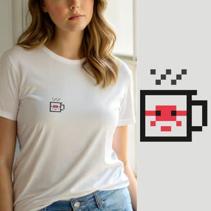 Pode incluir: Uma t-shirt branca de manga curta com um pequeno gráfico de pixel art centrado que representa uma caneca de café. A caneca está delineada a preto com uma faixa horizontal vermelha e um detalhe quadrado vermelho, com três marcas de vapor.