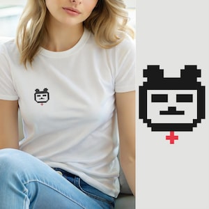 May include: A white short-sleeved t-shirt featuring a small, centred pixel art graphic on the chest. The graphic depicts a black and white square-shaped bear face with a small red cross symbol positioned directly below the chin of the bear.