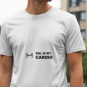 Może przedstawiać: Biała koszulka z czarnym napisem "SQL IS MY CARDIO". Koszulka ma okrągły dekolt i krótkie rękawy. Tekst jest wyśrodkowany na piersi. W tle widać ulicę i budynki.