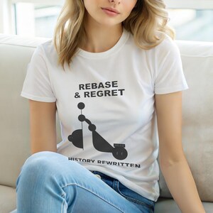 Puede incluir: Camiseta blanca con el texto "REBASE & REGRET" sobre un gráfico negro que se asemeja a un patinete. Debajo, el texto dice "HISTORY REWRITTEN". La camiseta está siendo usada.