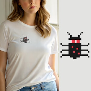 Könnte beinhalten: Ein weißes T-Shirt mit kurzen Ärmeln und einer kleinen Pixel-Art-Grafik auf der linken Brust. Die Grafik zeigt ein schwarzes Insekt mit roten Augen und roten Punkten auf dem Körper, dargestellt in einem blockartigen Digitalstil mit sechs Beinen und zwei Fühlern.