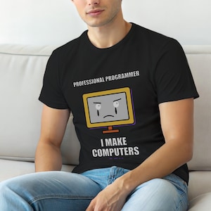 Op de afbeelding: Zwart T-shirt met de tekst "PROFESSIONAL PROGRAMMER" boven een cartooncomputer met een verdrietig gezicht. Daaronder staat de tekst "I MAKE COMPUTERS". Het T-shirt wordt door een persoon gedragen.