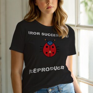 Op de afbeelding: Zwart T-shirt met een cartoon lieveheersbeestje ontwerp en de tekst "ERROR SUCCESSFULLY REPRODUCED" in het wit. Het lieveheersbeestje is rood met zwarte stippen en een blauwe kop. Het shirt heeft korte mouwen.