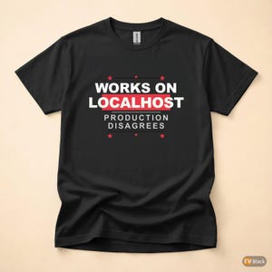 Può includere: T-shirt nera con la scritta "WORKS ON LOCALHOST" in bianco e rosso, con la frase "PRODUCTION DISAGREES" sotto. Grafiche di stelle rosse sono sopra e sotto il testo.