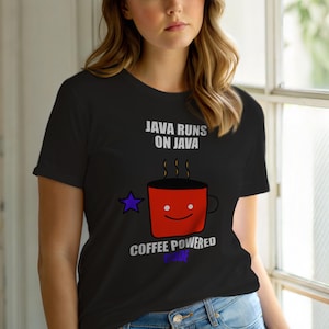 Op de afbeelding: Zwart T-shirt met de tekst "JAVA RUNS ON JAVA" boven een rode koffiemok met een lachend gezicht. Onder de mok staat de tekst "COFFEE POWERED CODE". Links van de mok staat een paarse ster.