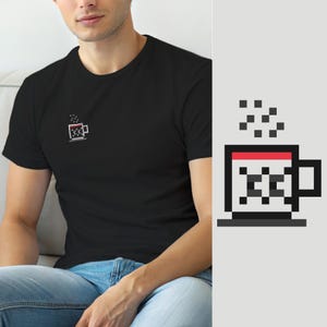 Puede incluir: Una camiseta negra de manga corta con un pequeño gráfico de pixel art en el pecho izquierdo. El gráfico muestra una taza blanca con borde rojo, asa negra y un patrón de cuadros negros en el lateral, con vapor gris pixelado elevándose arriba.