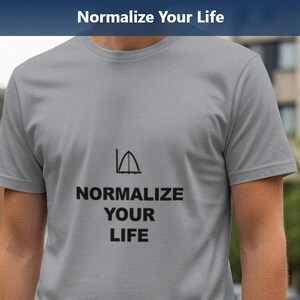 May include: A gray t-shirt with the text "Normalize Your Life" printed in black. The shirt features a graphic above the text. The image is taken outdoors with a blurred background of buildings and trees.