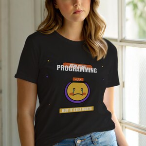 Op de afbeelding: Zwart T-shirt met de tekst "HTML IS NOT PROGRAMMING BUT IT STILL HURTS". Het shirt heeft een verdrietige gezichtsafbeelding met een paarse omtrek en een codeersymbool. Het shirt heeft korte mouwen.