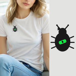 Op de afbeelding: Een wit T-shirt met korte mouwen en een zwarte afbeelding van een kever met een felgroene pleister op de rug. De kever heeft zes poten en twee voelsprieten, waarbij de groene pleister in het midden van het ronde lichaam is geplaatst.