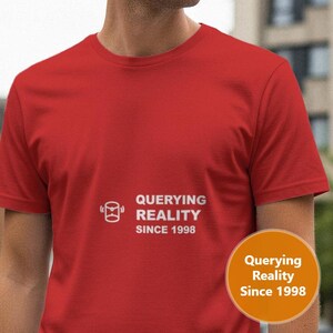 May include: A red crew neck t-shirt with the text "QUERYING REALITY SINCE 1998" printed in white. The shirt has short sleeves and a round neckline. A circular orange sticker with the same text is visible.