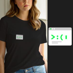 Op de afbeelding: Een zwart t-shirt met korte mouwen en een kleine afbeelding op de borst. De afbeelding toont een lichtgrijs computerterminalvenster met een groene opdrachtprompt die de tekst >:( weergeeft, gevolgd door een knipperende verticale cursor.