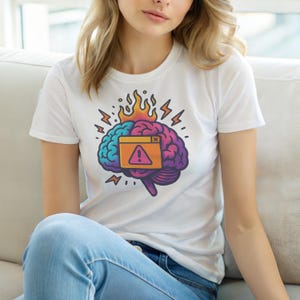 May include: White t-shirt featuring a colorful brain graphic with flames, lightning bolts, and an alert symbol. The brain illustration is in shades of blue, purple, and orange. The shirt is made of a soft material.