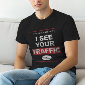 Op de afbeelding: Zwart T-shirt met de tekst "I SEE YOUR TRAFFIC" in wit en rood. Het shirt heeft ook de tekst "[ PACKET SNIFFER ]" boven de hoofdtekst en een afbeelding van een cd. Het shirt wordt door een persoon gedragen.