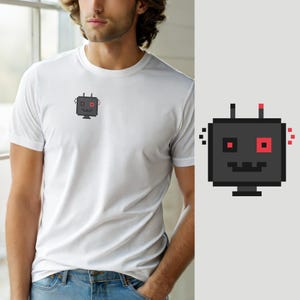 Könnte beinhalten: Ein weißes Kurzarm-T-Shirt mit einer kleinen, zentrierten Pixel-Art-Grafik auf der Brust. Die Grafik zeigt einen quadratischen, dunkelgrauen Roboterkopf mit zwei Antennen, einem gezackten Mund und zwei markanten roten Quadrat-Augen.