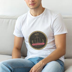 Puede incluir: Camiseta blanca con un diseño gráfico circular. El diseño incluye la palabra "GIT", un violín estilizado y la frase "IT WORKS NOW" en rosa. El diseño tiene un fondo oscuro y un borde dorado.