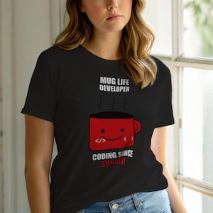 Op de afbeelding: Zwart T-shirt met de tekst "MUG LIFE DEVELOPER" boven een rode mok-afbeelding. De mok heeft een lachend gezicht en codeersymbolen. Daaronder staat "CODING SINCE SUNRISE".