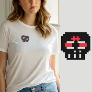 Op de afbeelding: Een wit T-shirt met korte mouwen en een kleine pixel art schedelafbeelding op de linkerborst. Het schedelontwerp is uitgevoerd in zwart, wit en rood, met een blokkerige, digitale esthetiek die doet denken aan vroege 8-bit videogames.