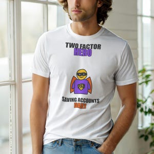 Op de afbeelding: Wit t-shirt met de tekst "TWO FACTOR HERO" in zwart en paars. Daaronder staat een cartoon superheld met een geel masker, paars shirt en oranje cape. De tekst "SAVING ACCOUNTS DAILY" staat eronder.