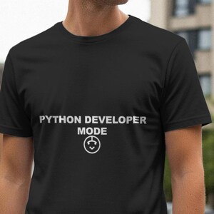 Op de afbeelding: Zwart T-shirt met de witte tekst "PYTHON DEVELOPER MODE" en een cirkelvormige afbeelding. Het shirt met korte mouwen lijkt van een zacht materiaal te zijn. De achtergrond toont een vage stedelijke omgeving.